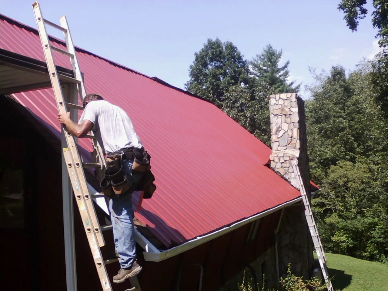 Roofer on a ladder beside a red metal roof and stone chimney — Soffit & Fascia Repair by Roofing Brothers in Susquehanna